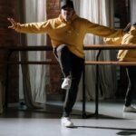Energetic hip hop dancer in a yellow sweater striking a pose indoors with brick wall backdrop.