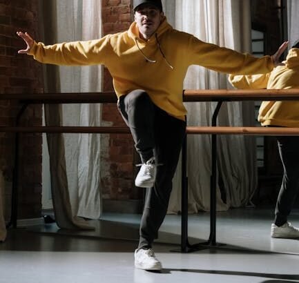 Energetic hip hop dancer in a yellow sweater striking a pose indoors with brick wall backdrop.