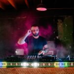 A DJ performs with vibrant light effects during a music event in a nightclub setting.