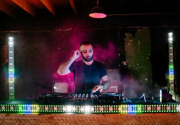 A DJ performs with vibrant light effects during a music event in a nightclub setting.